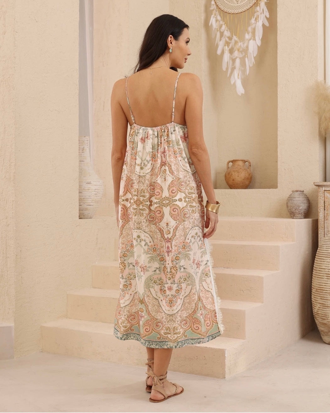 A woman wearing a sleeveless midi slip dress with thin straps and a paisley pattern in beige, pink, and green, shown from the back in a warm-toned indoor setting with steps and decorative vases.