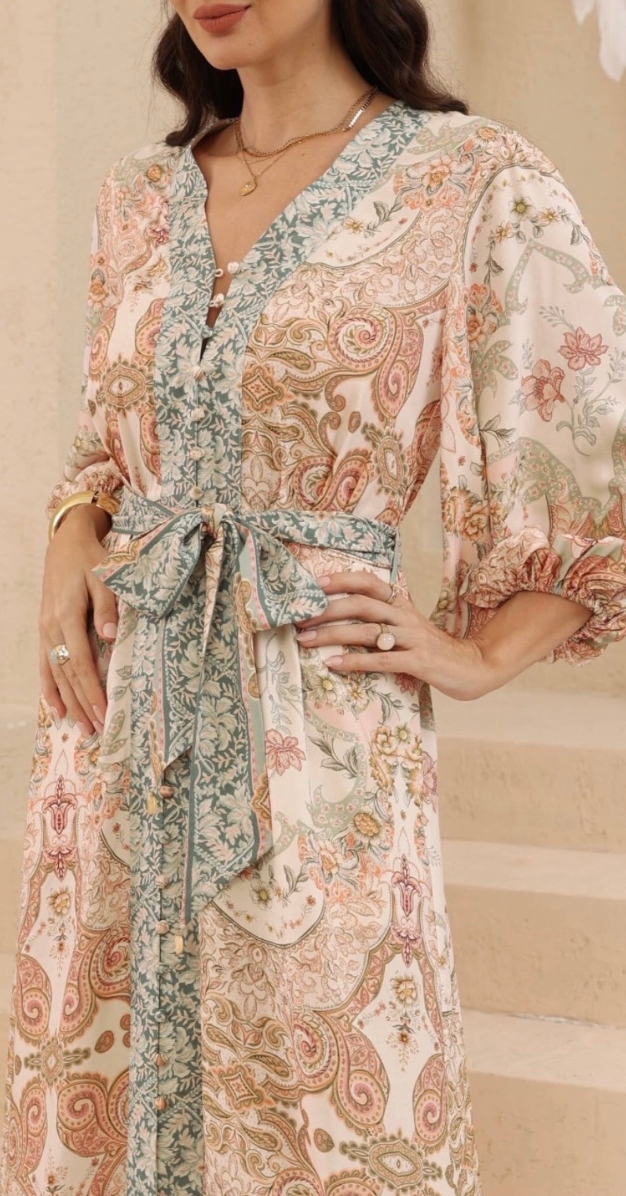 A woman wearing a long-sleeve V-neck wrap dress with a tie waist and a paisley floral pattern in soft beige, dusty pink, and sage green tones.