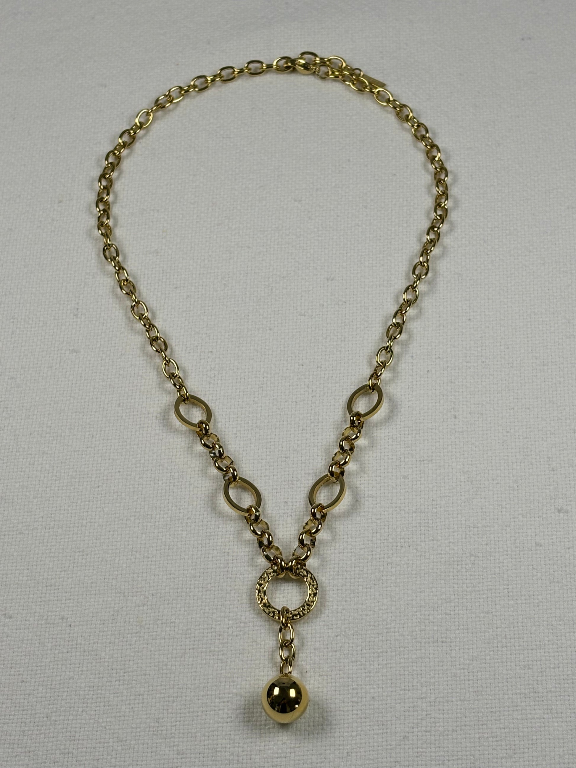 A gold-toned chain necklace with oval links, a textured ring connector, and a dangling polished bead pendant, displayed on a light fabric background.