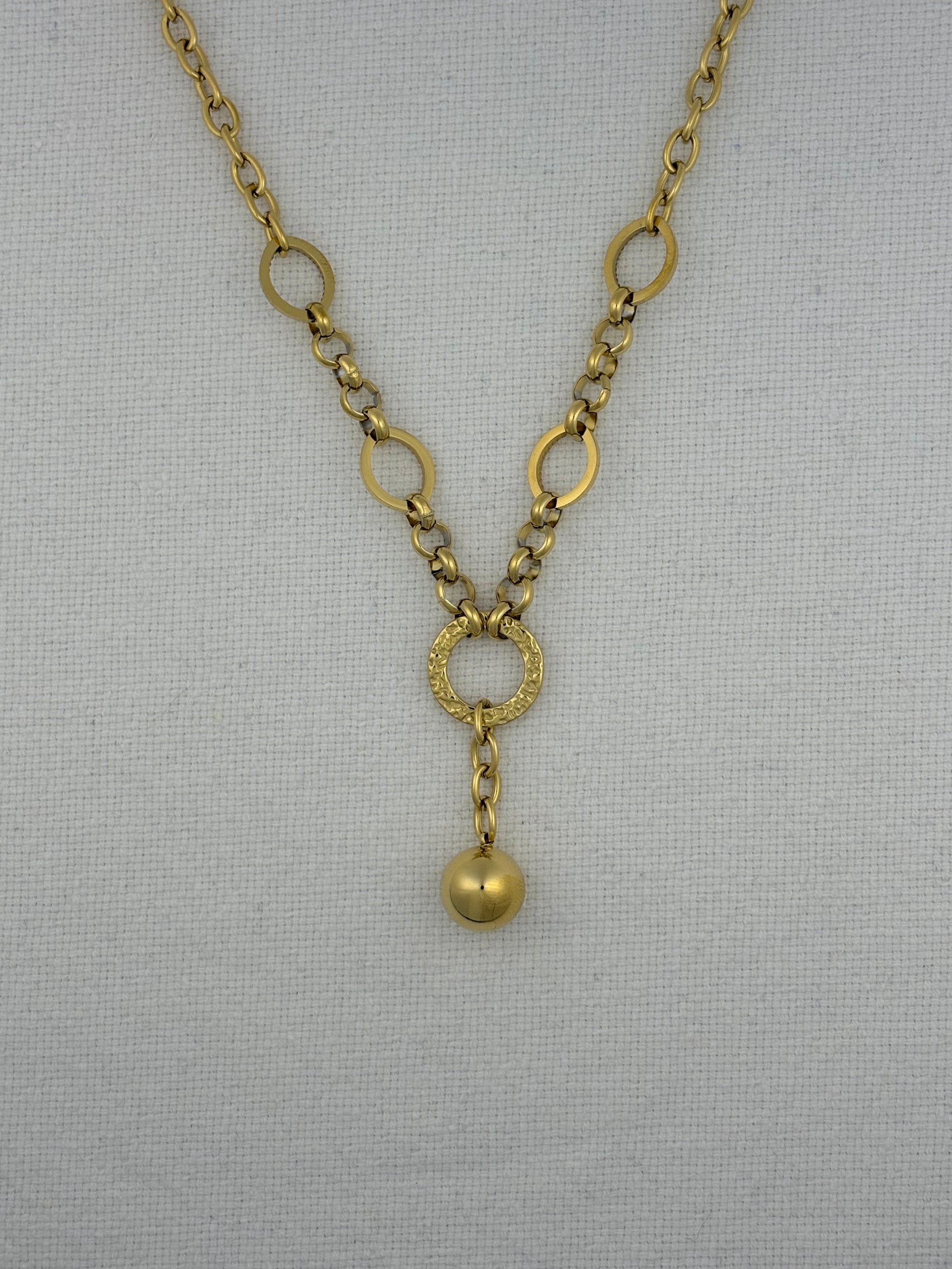 A gold-toned chain necklace with alternating large and small circular links, a textured ring connector, and a dangling polished bead pendant, displayed on a light gray fabric background.