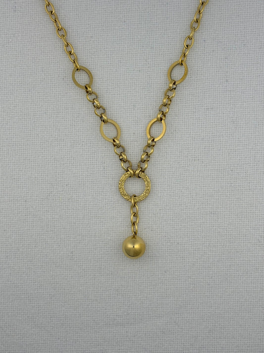 A gold-toned chain necklace with alternating large and small circular links, a textured ring connector, and a dangling polished bead pendant, displayed on a light gray fabric background.