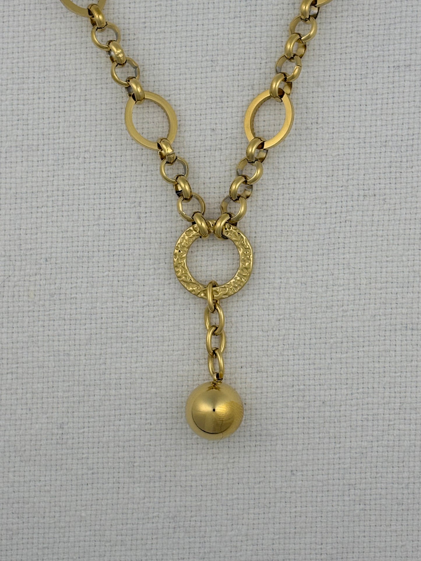 A gold-toned chain necklace featuring oval links, a textured circular ring connector, and a dangling polished sphere charm, displayed on a white textured fabric background.