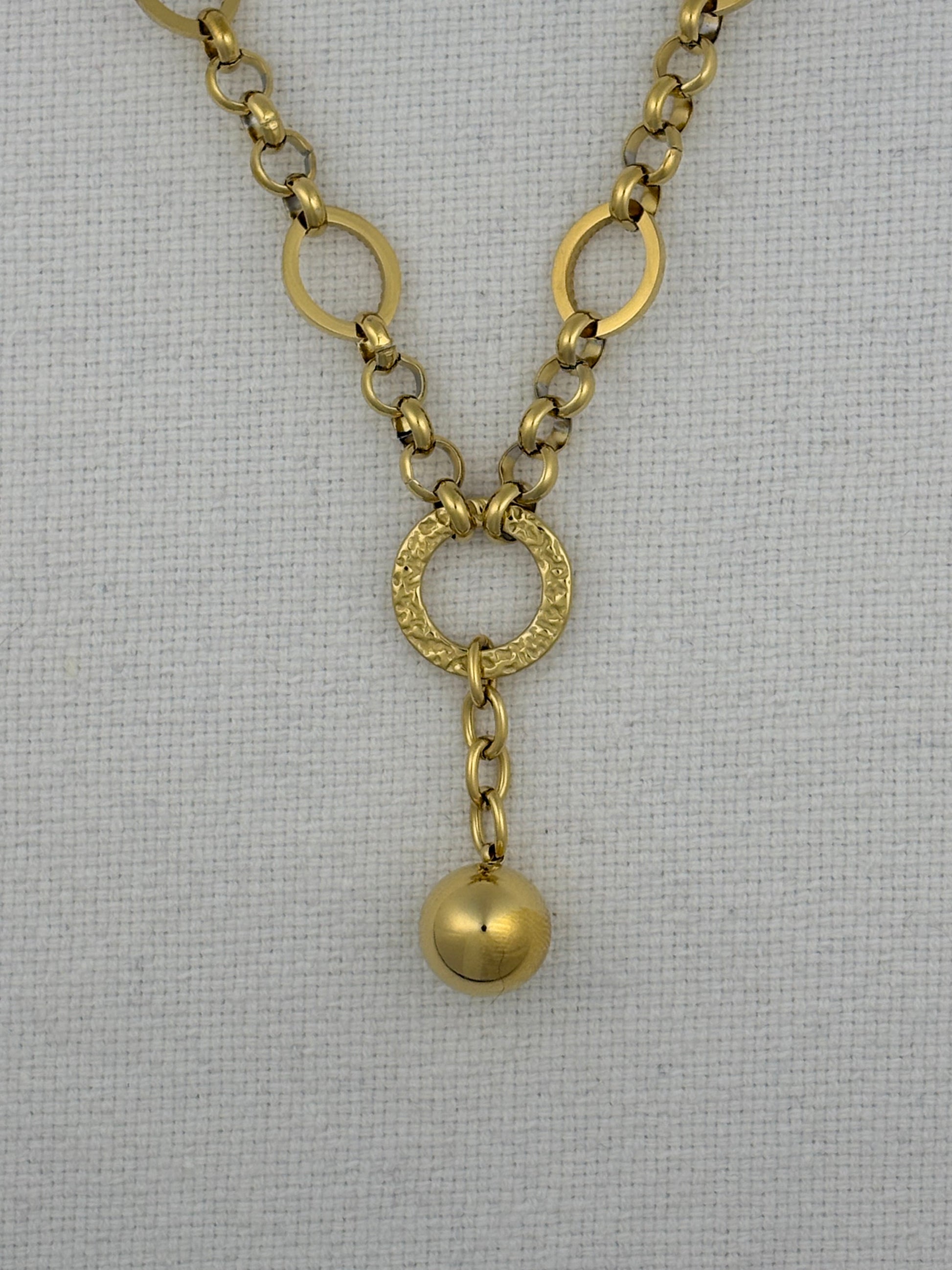 A gold-toned chain necklace featuring oval links, a textured circular ring connector, and a dangling polished sphere charm, displayed on a white textured fabric background.
