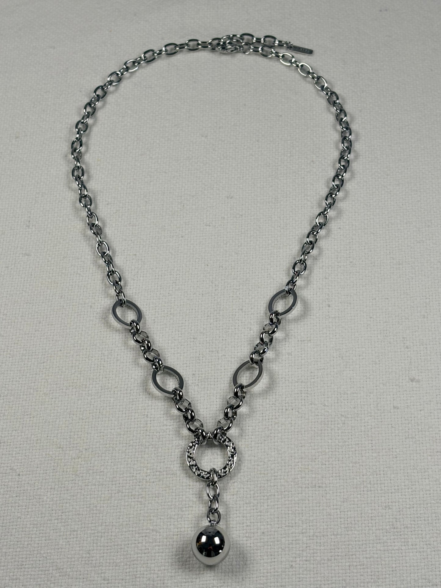 A silver-toned chain necklace with alternating oval and circular links, a textured ring connector, and a polished spherical pendant, displayed on a light beige fabric background.