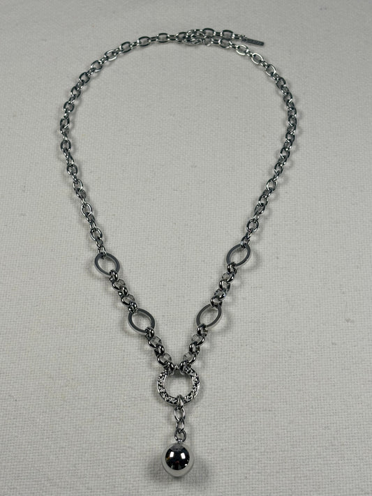 A silver-toned chain necklace with alternating oval and circular links, a textured ring connector, and a polished spherical pendant, displayed on a light beige fabric background.