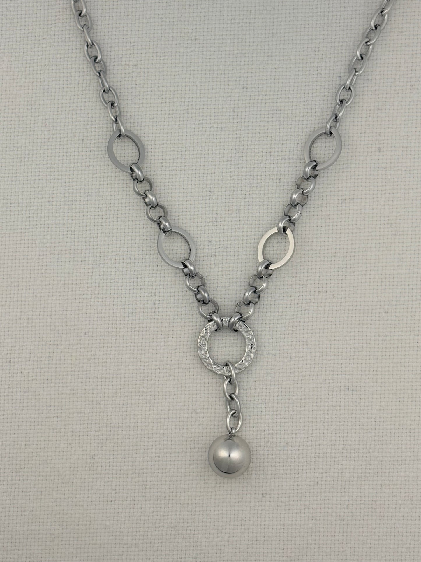 A silver-toned necklace with a textured chain, circular link details, a pavé-set ring, and a dangling pearl-like bead, displayed on a neutral textured fabric background.