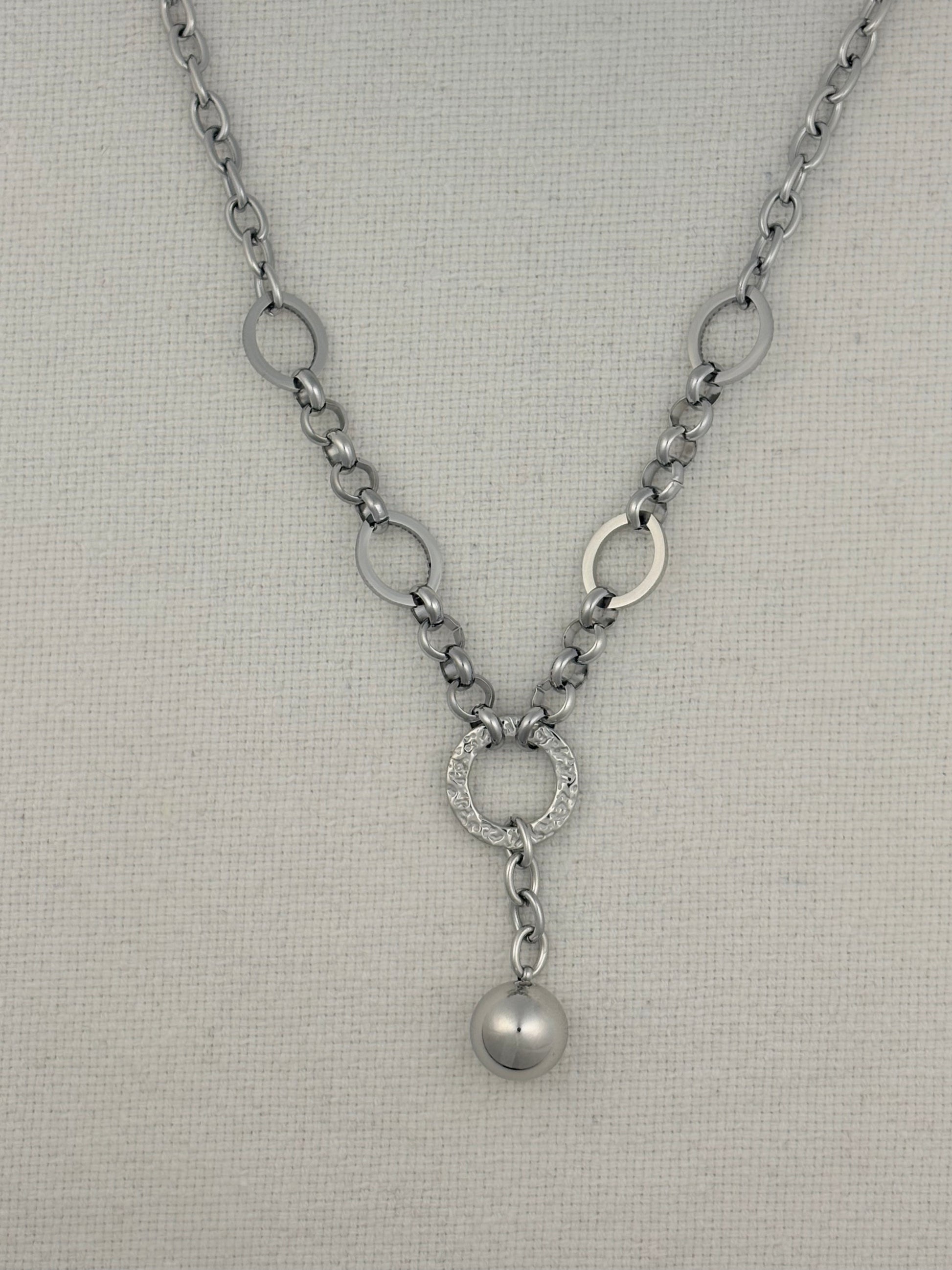 A silver-toned necklace with a textured chain, circular link details, a pavé-set ring, and a dangling pearl-like bead, displayed on a neutral textured fabric background.