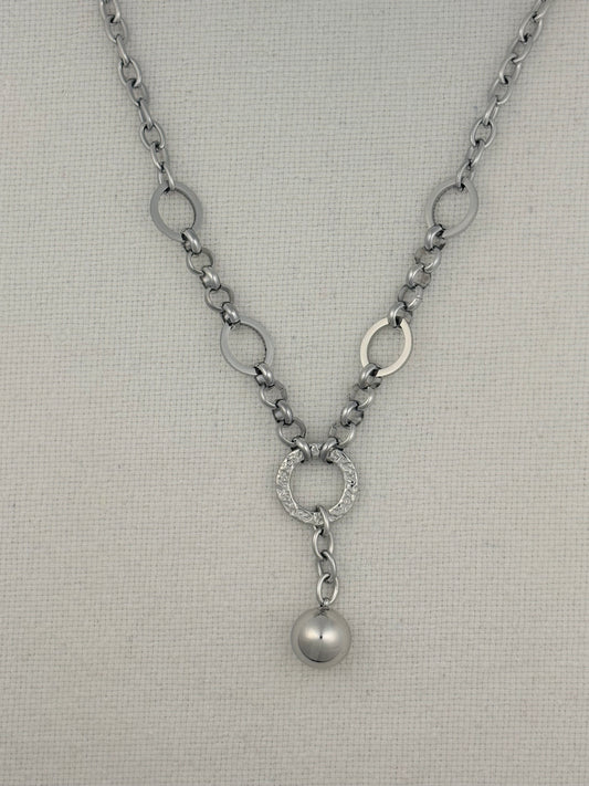 A silver-toned necklace with a textured chain, circular link details, a pavé-set ring, and a dangling pearl-like bead, displayed on a neutral textured fabric background.