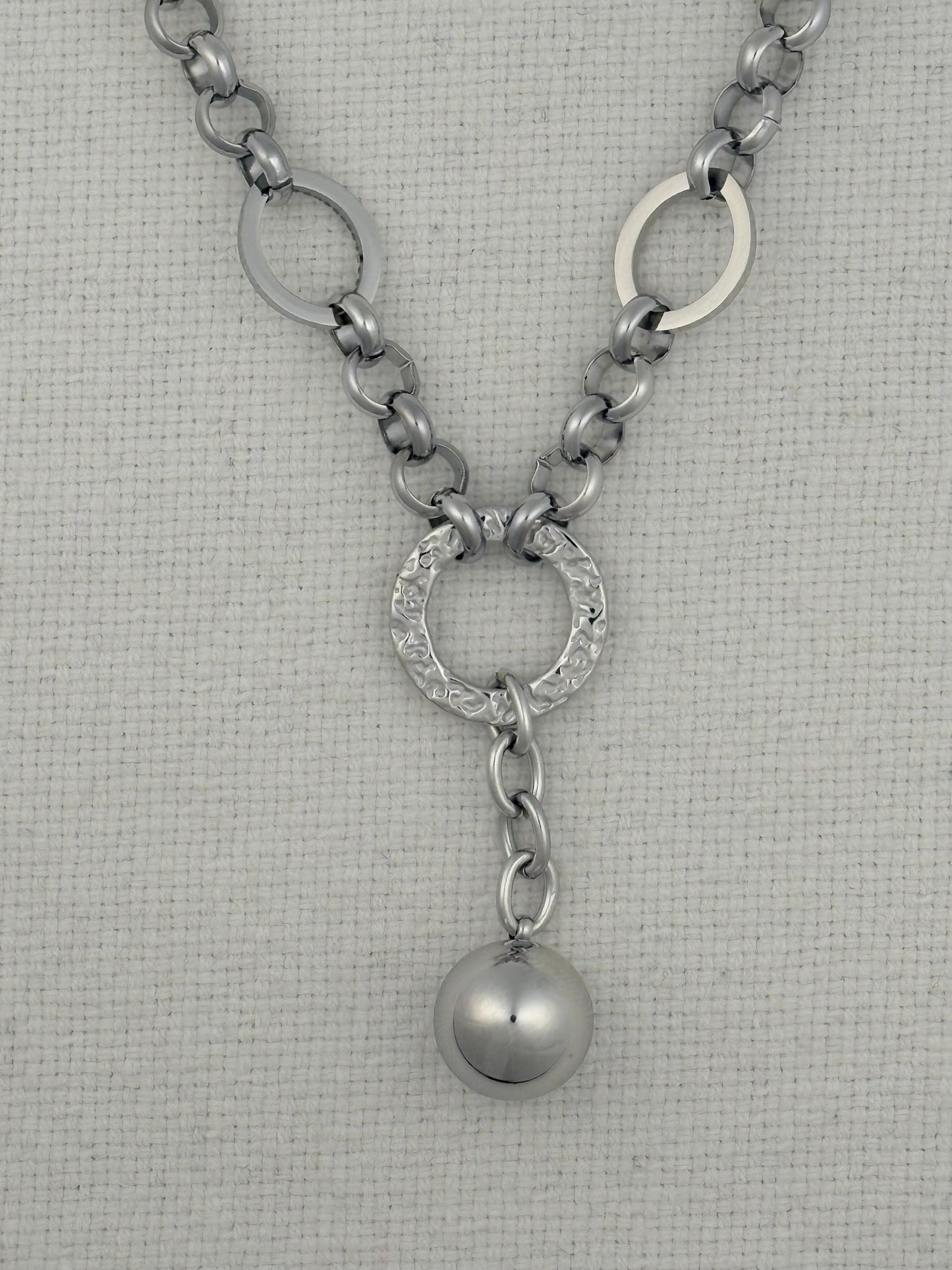 A silver-toned chain necklace featuring a Y-shaped design with three interlocking circular links at the top, a textured oval center link, and a dangling polished sphere charm, displayed on a textured off-white fabric background.