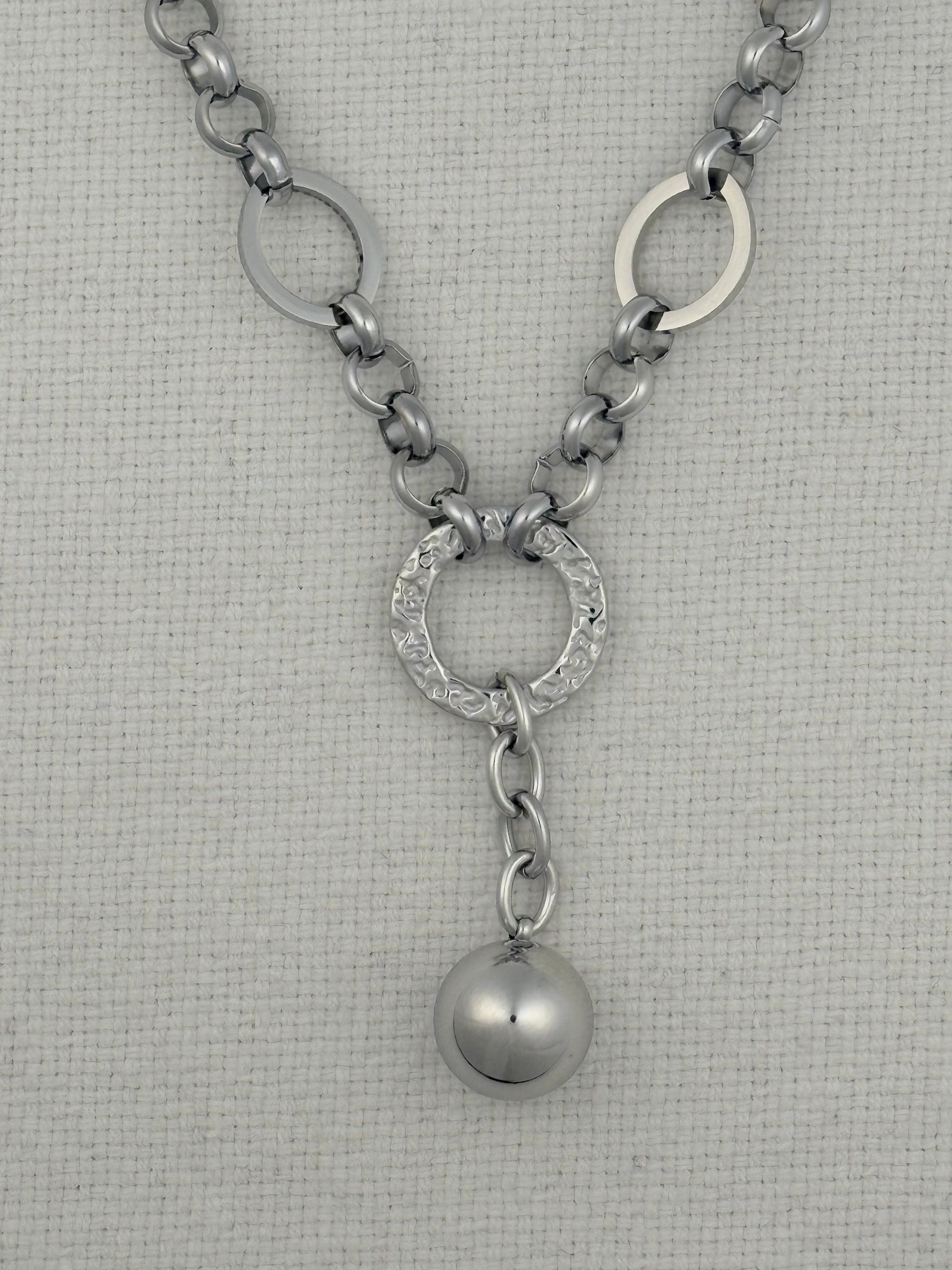 A silver-toned chain necklace featuring a Y-shaped design with three interlocking circular links at the top, a textured oval center link, and a dangling polished sphere charm, displayed on a textured off-white fabric background.