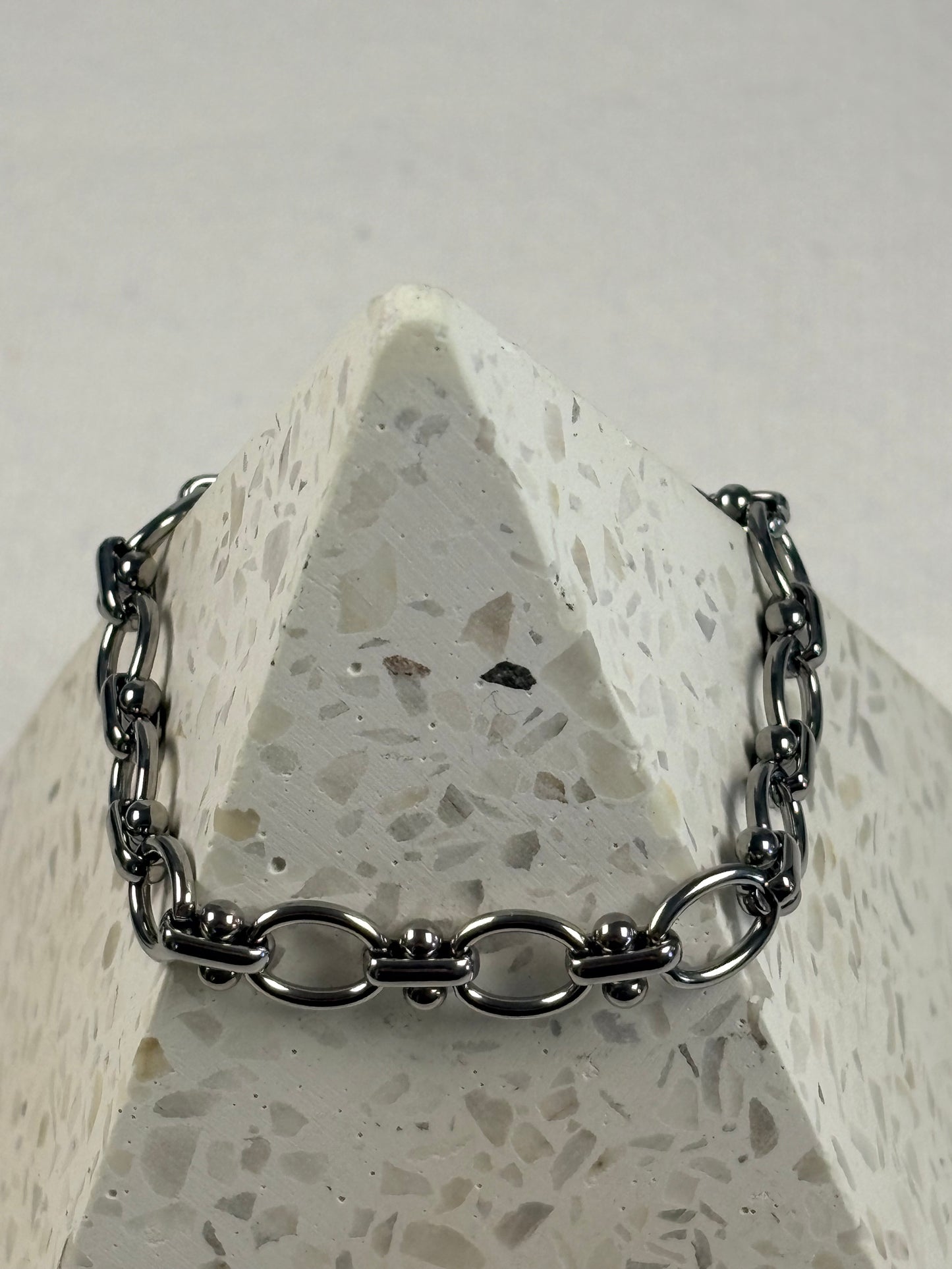 A silver chain bracelet with distinctive oval links displayed on a white speckled stone pyramid stand.
