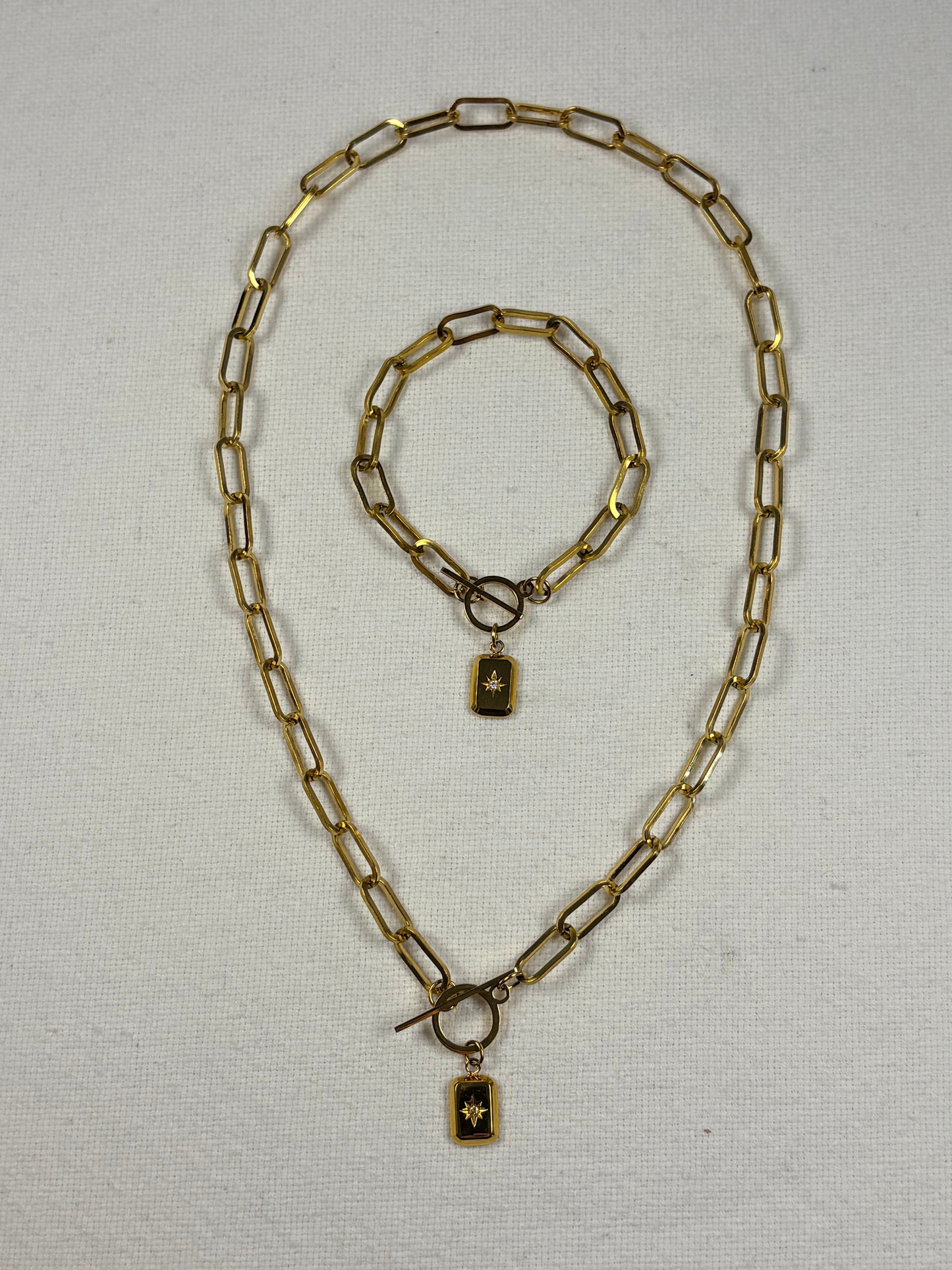 A gold-toned metal jewelry set consisting of a chain necklace and a matching bracelet, laid flat on a textured off-white fabric background. 