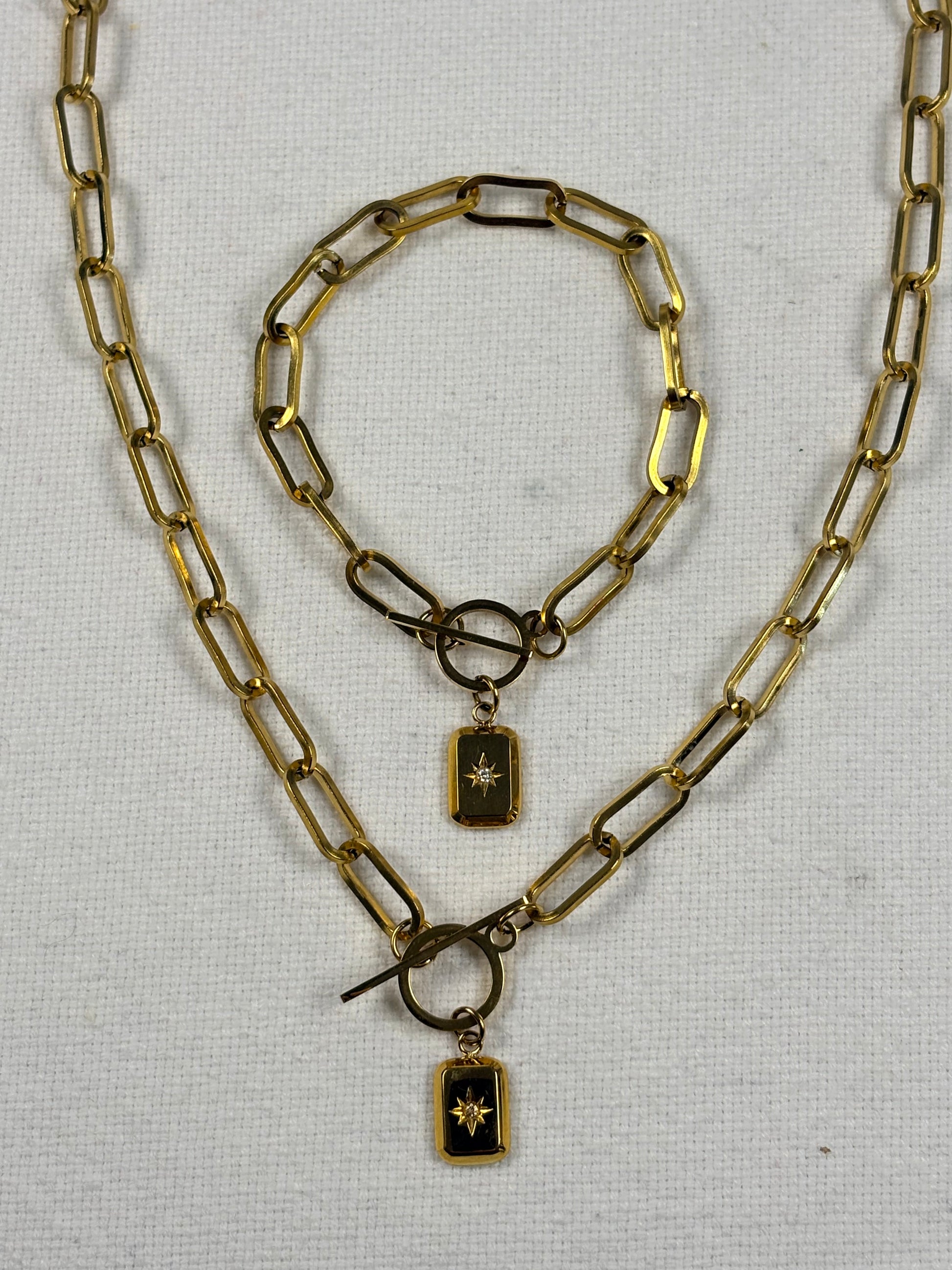 A gold-toned chain necklace and matching bracelet with rectangular links and small rectangular pendants featuring black centers with gold starburst details, displayed on a white textured fabric background.