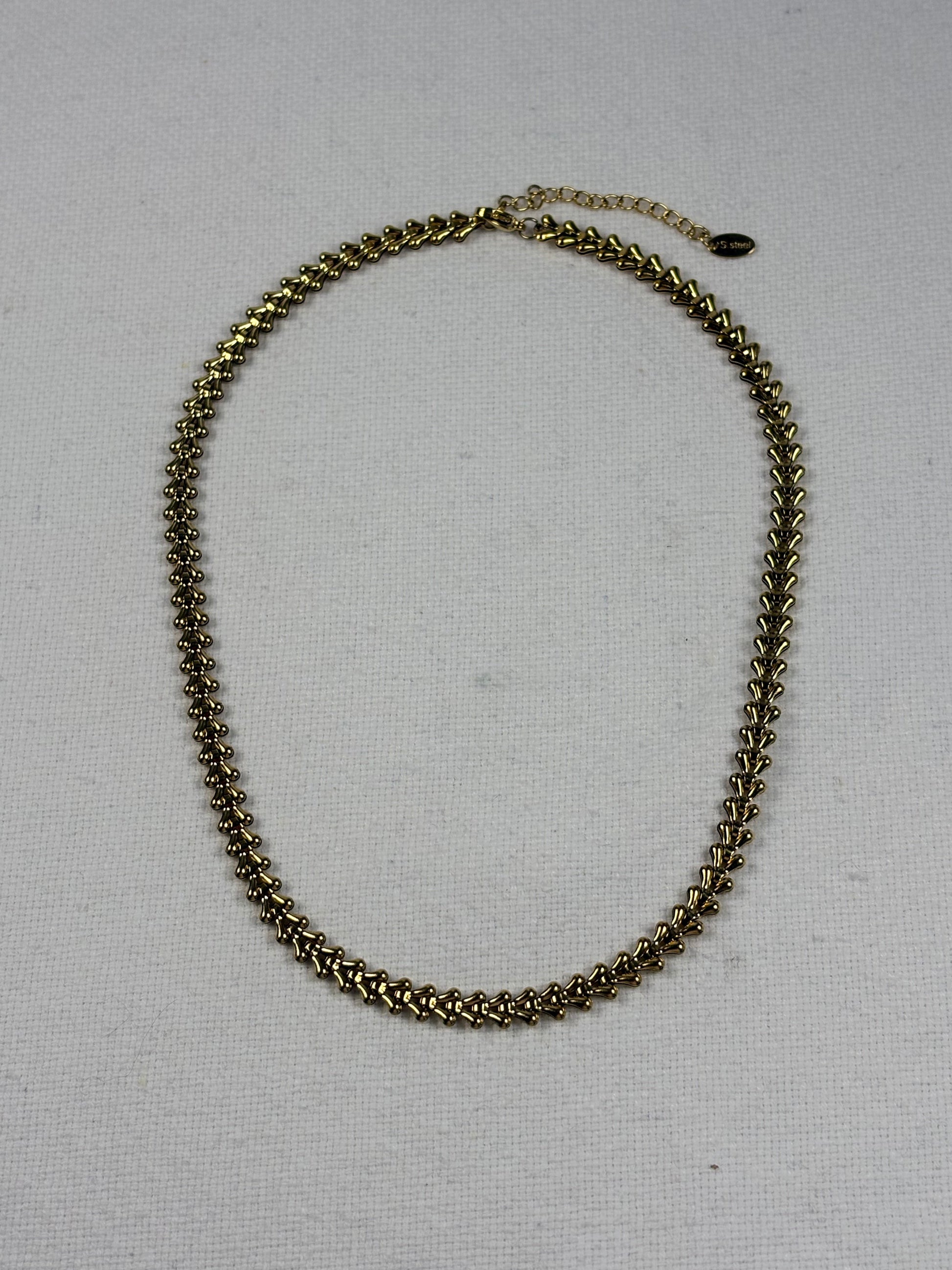 A gold-toned chain necklace with a textured, woven link design and an extender chain with a small dark bead, displayed on a light beige fabric background.