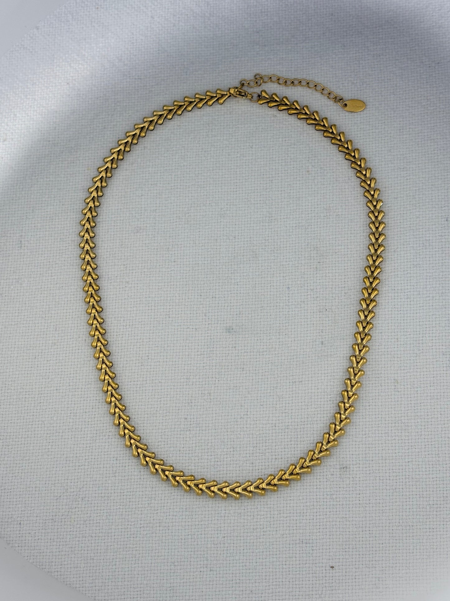 A gold-toned chain necklace with a textured, woven chevron pattern, displayed on a light gray textured fabric background.
