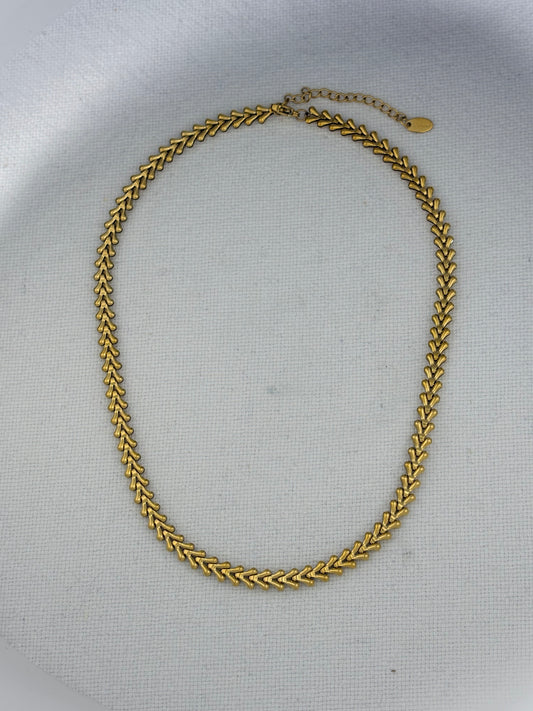 A gold-toned chain necklace with a textured, woven chevron pattern, displayed on a light gray textured fabric background.