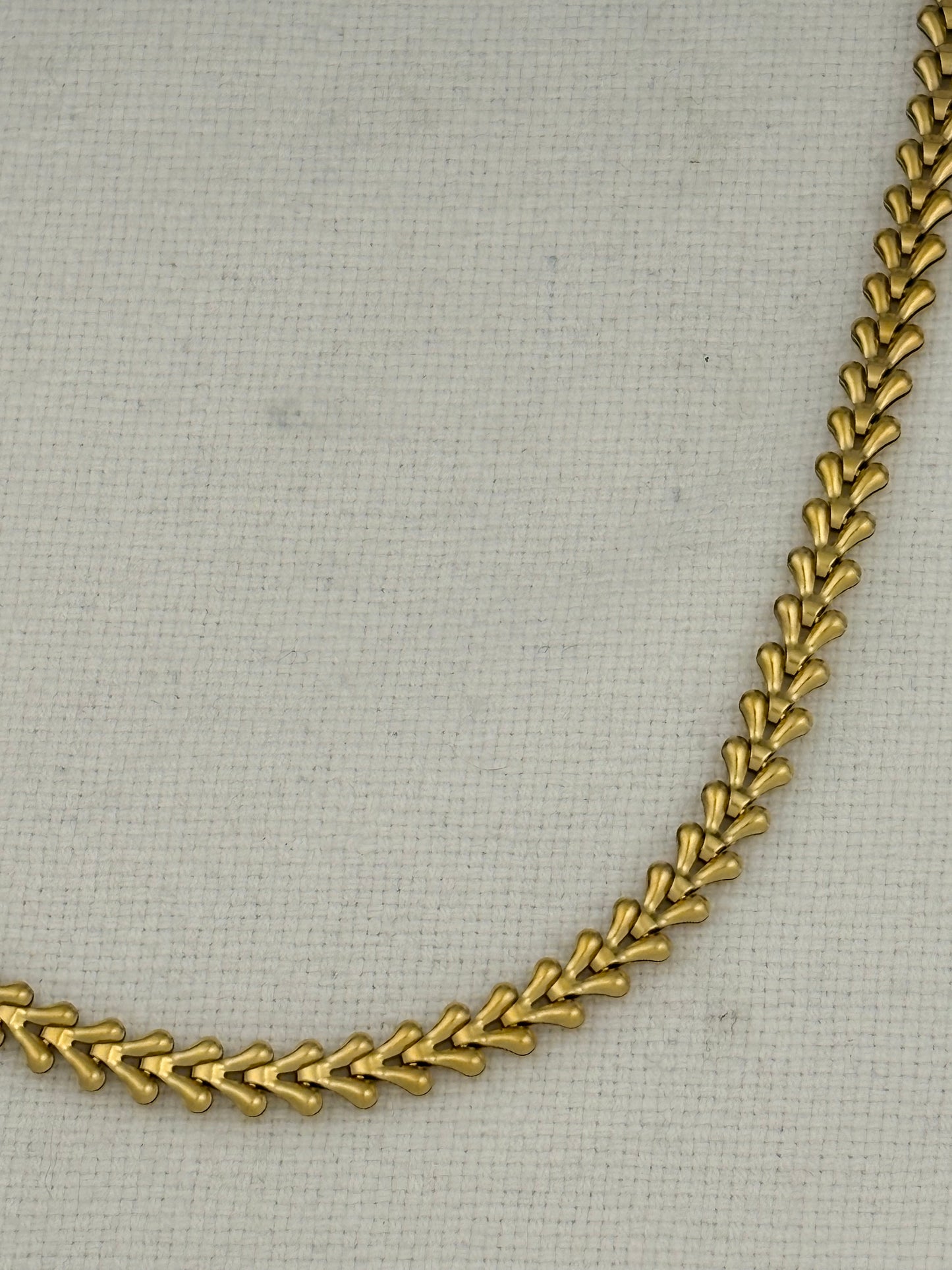 A close-up of a gold chain necklace with a repeating leaf or chevron pattern, laid on a light beige textured fabric background.