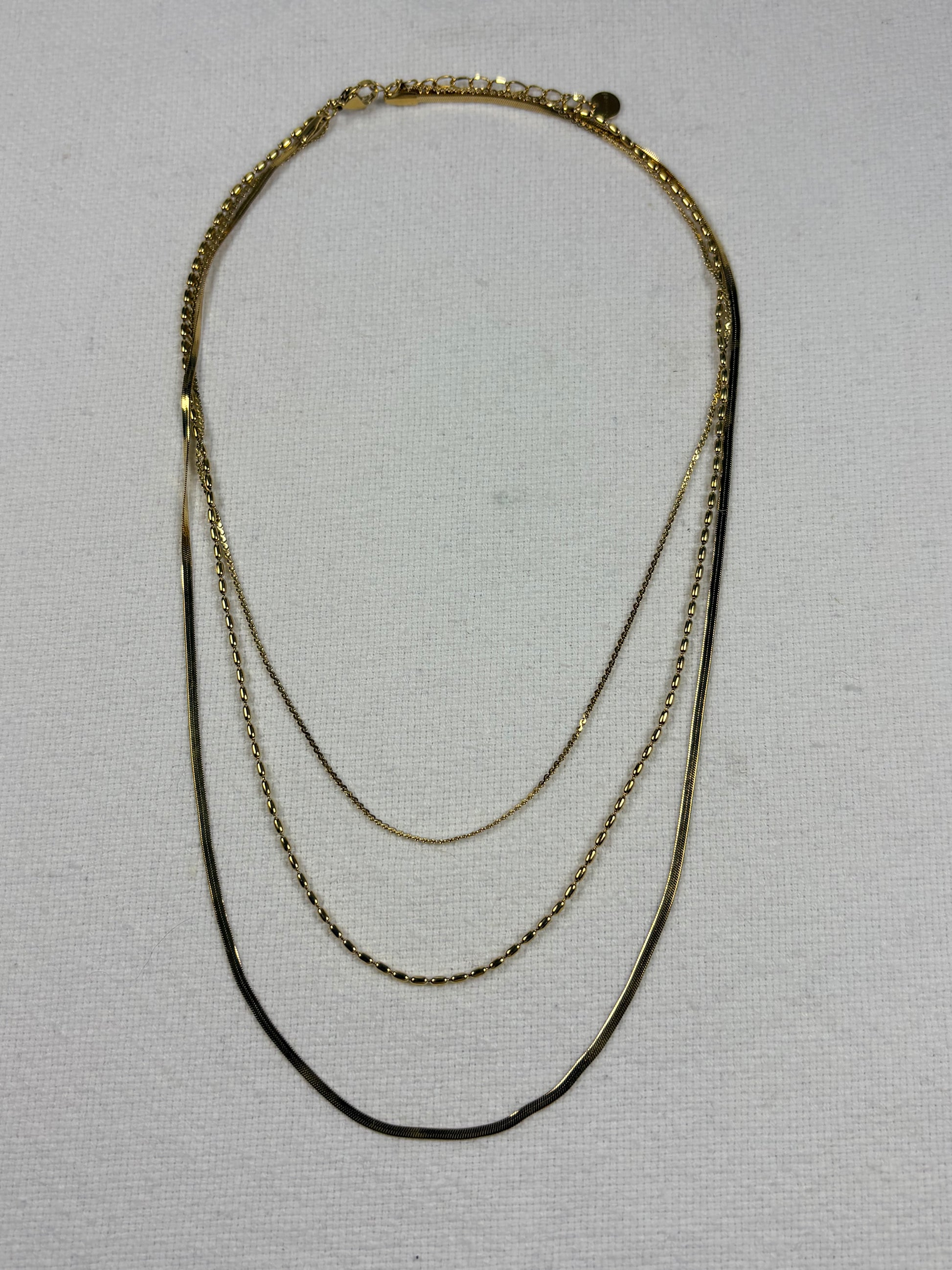 A three-layer gold chain necklace with varying chain widths, displayed on a textured off-white fabric background.