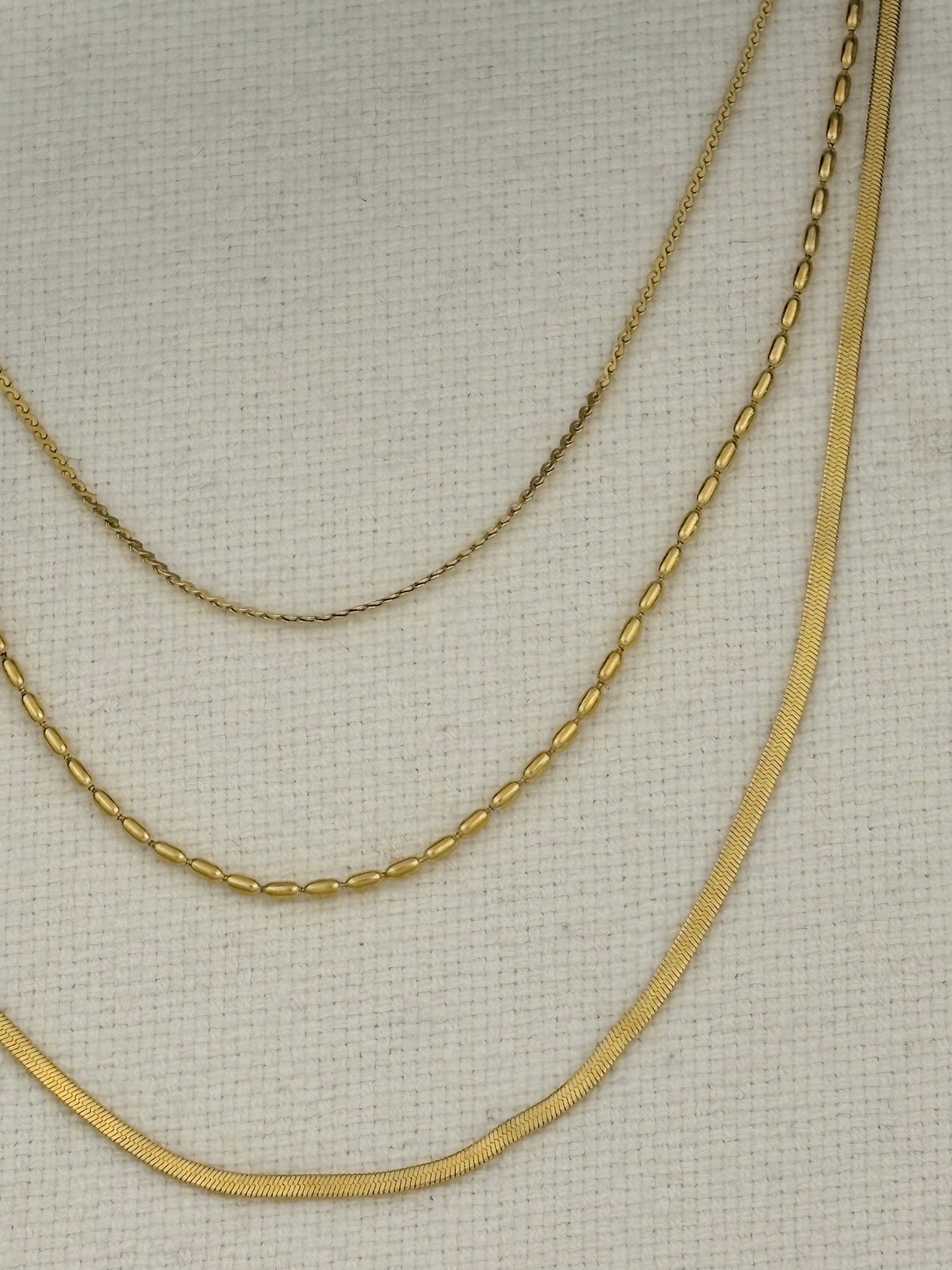 Three gold-tone chains of different widths and textures draped on a light beige textured fabric background.