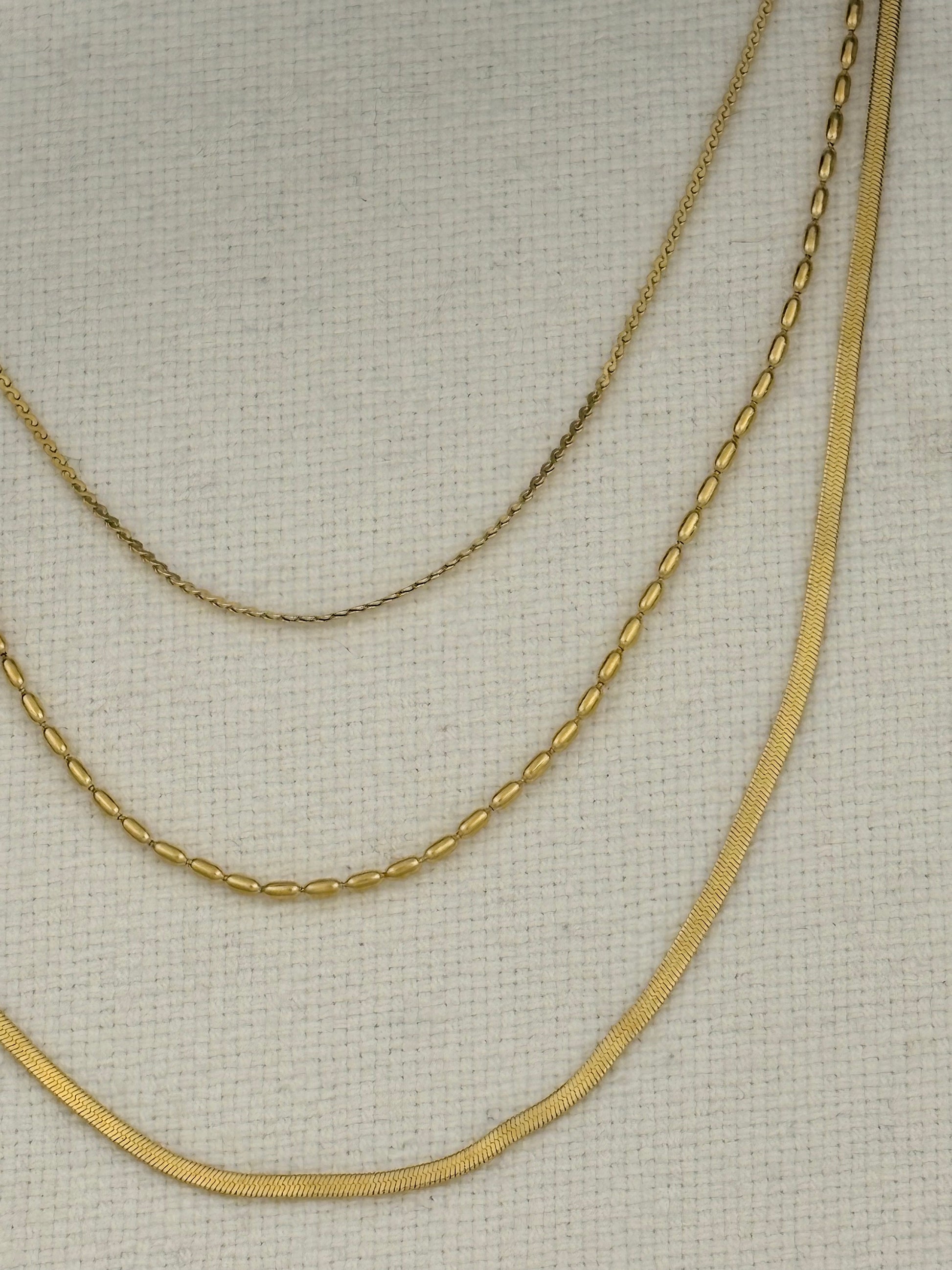 Three gold-tone chains of different widths and textures draped on a light beige textured fabric background.