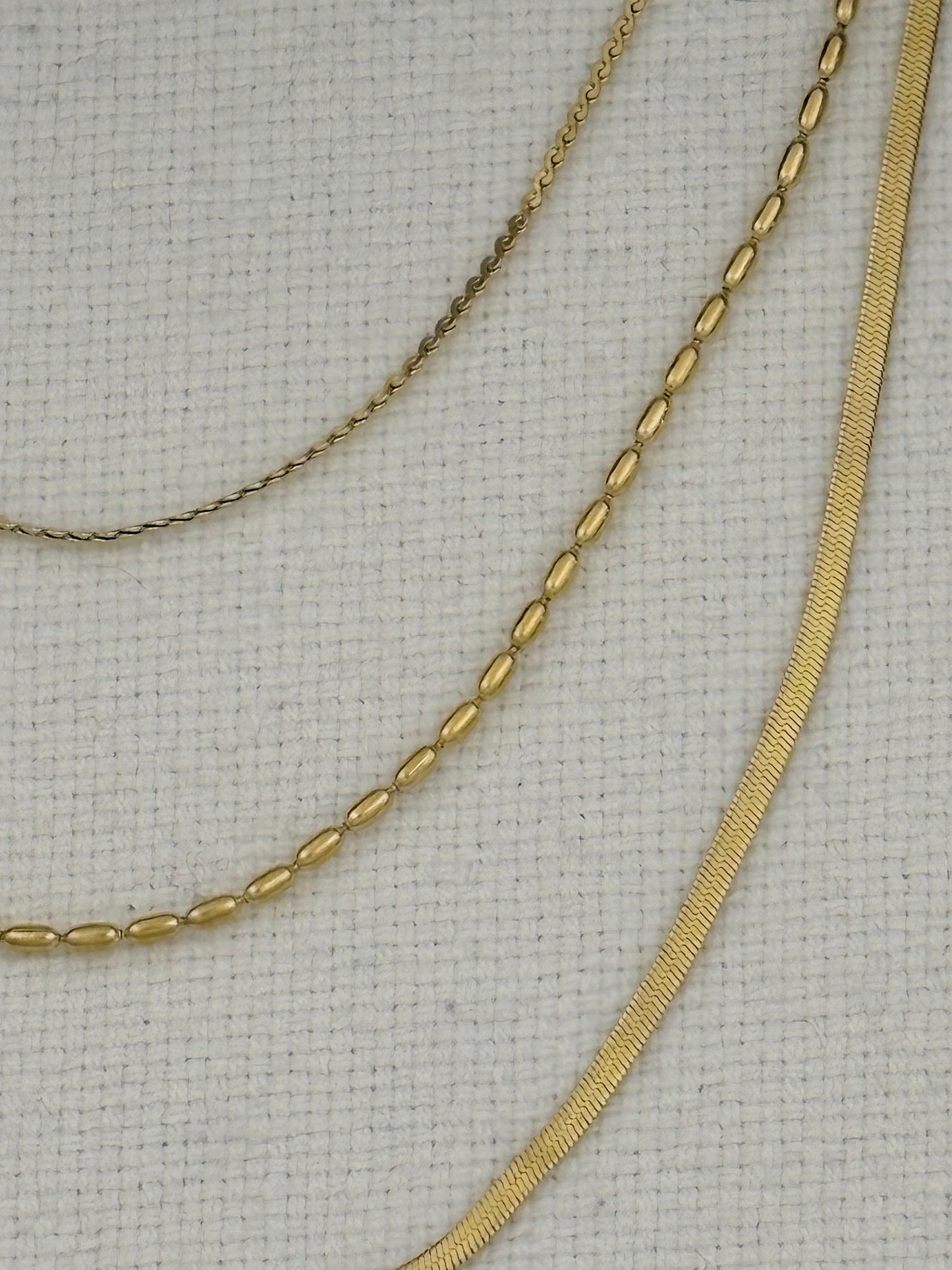 Three gold-tone chains displayed on a textured off-white fabric background: a delicate cable chain, a flat curb chain, and a tennis-style oval chain.