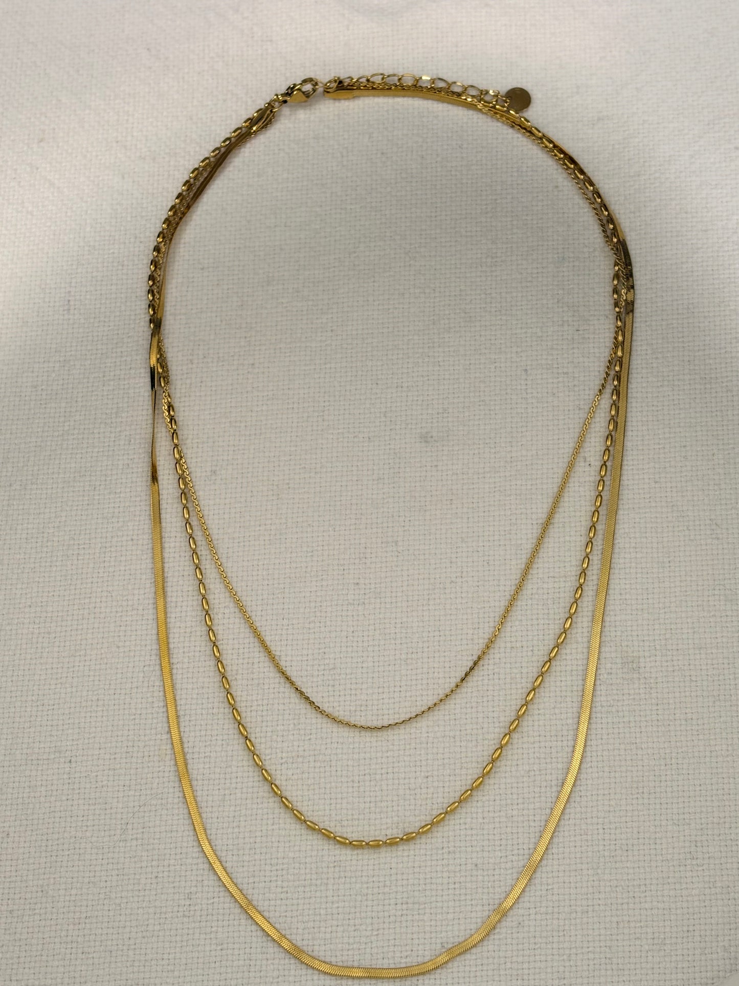 A gold multi-layer necklace featuring three distinct chain styles: a thick braided chain, a delicate cable chain, and a thin snake chain, displayed on a textured off-white fabric background.