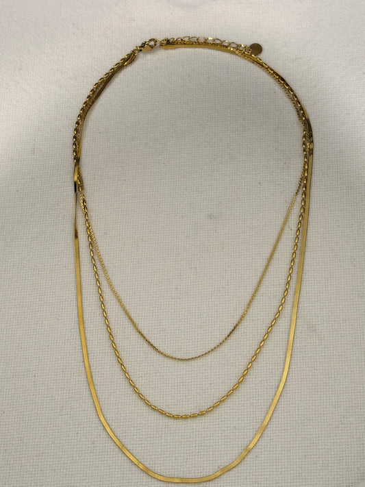 A gold multi-layer necklace featuring three distinct chain styles: a thick braided chain, a delicate cable chain, and a thin snake chain, displayed on a textured off-white fabric background.