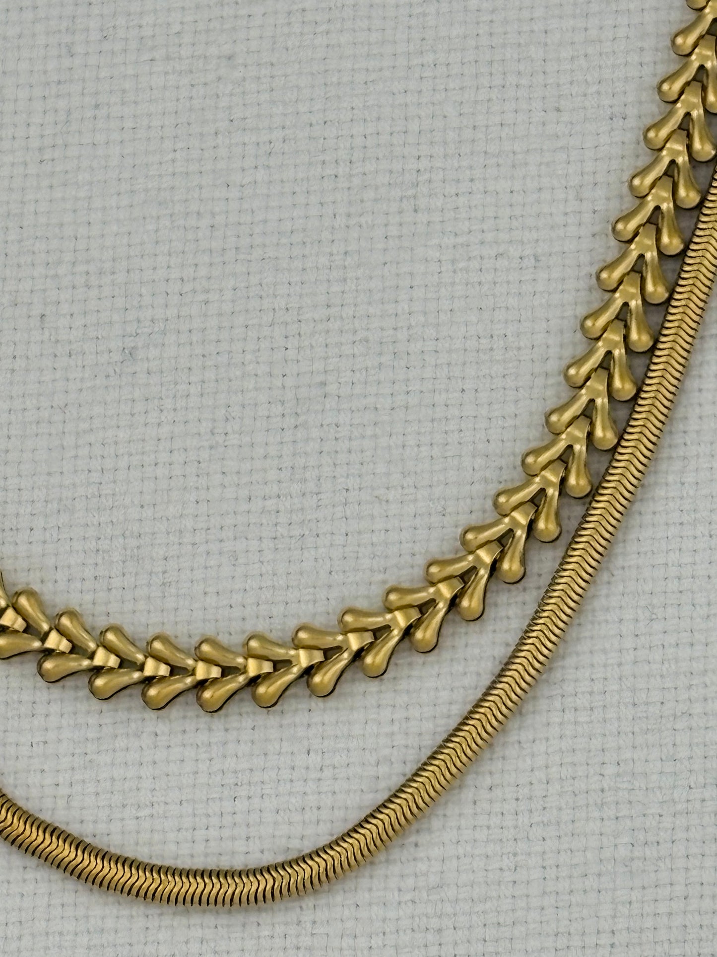 A close-up of a gold-toned chain necklace draped over a light gray textured fabric background, showing two linked chain styles.