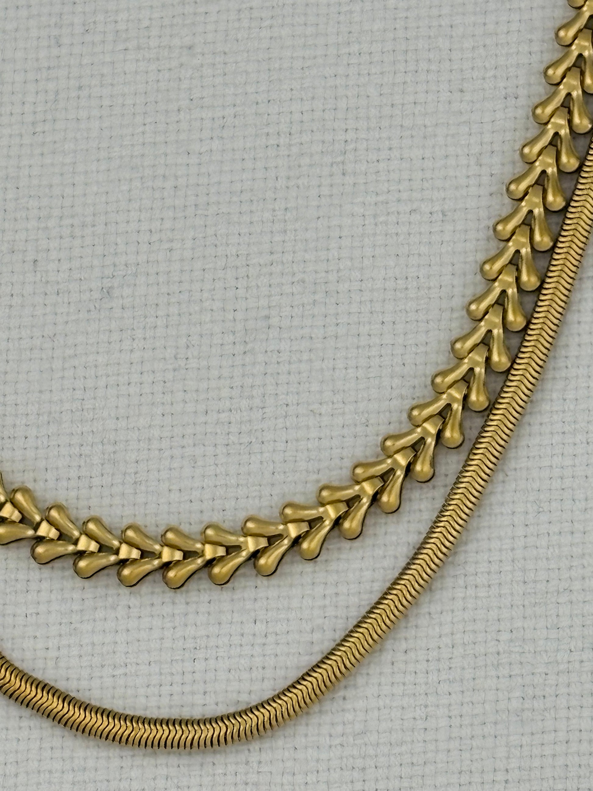 A close-up of a gold-toned chain necklace draped over a light gray textured fabric background, showing two linked chain styles.