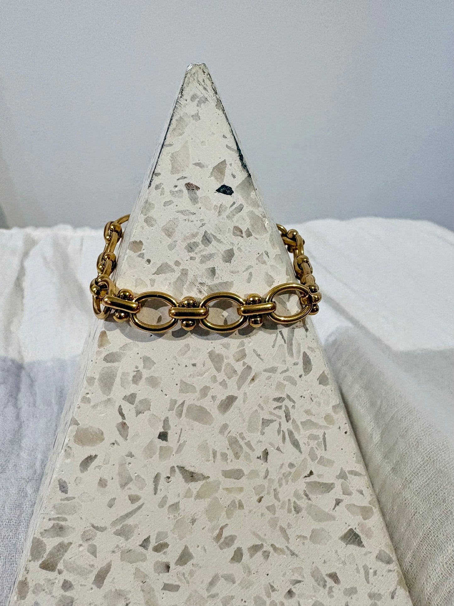 A gold-toned metal chain bracelet with interlocking oval links, displayed on a white terrazzo cone stand against a light gray background.