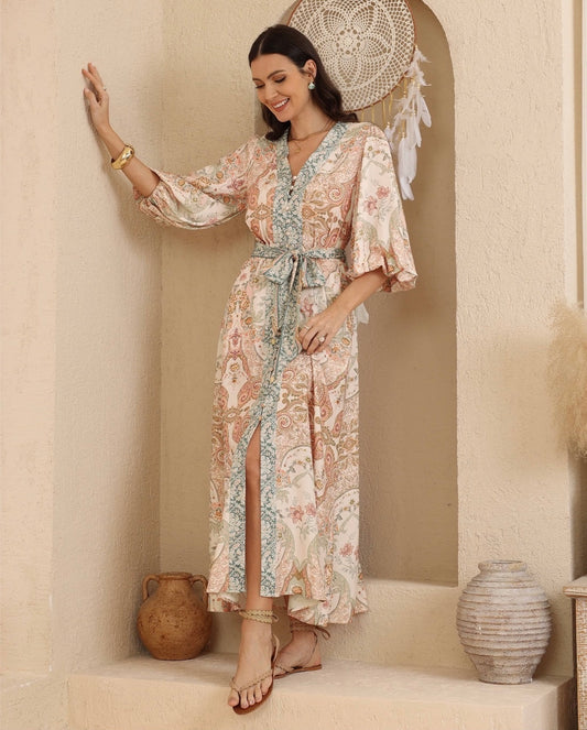 A woman models a bohemian-style maxi dress with a beige, pink, and teal paisley pattern, standing in a warm, neutral-toned room with decorative pottery and a dreamcatcher.