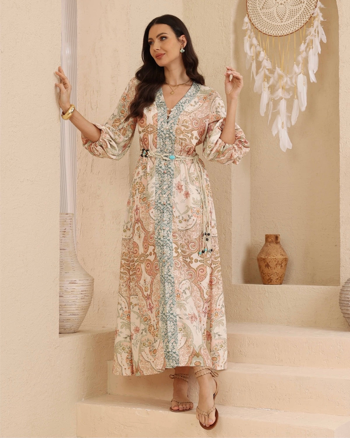 A woman models a long-sleeved maxi dress with a beige, pink, and blue paisley pattern, standing on beige stone steps in a bohemian-style room decorated with a hanging dreamcatcher and vases.