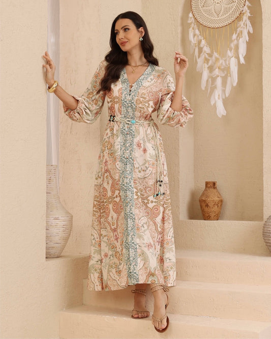 A woman models a long-sleeved maxi dress with a beige, pink, and blue paisley pattern, standing on beige stone steps in a bohemian-style room decorated with a hanging dreamcatcher and vases.