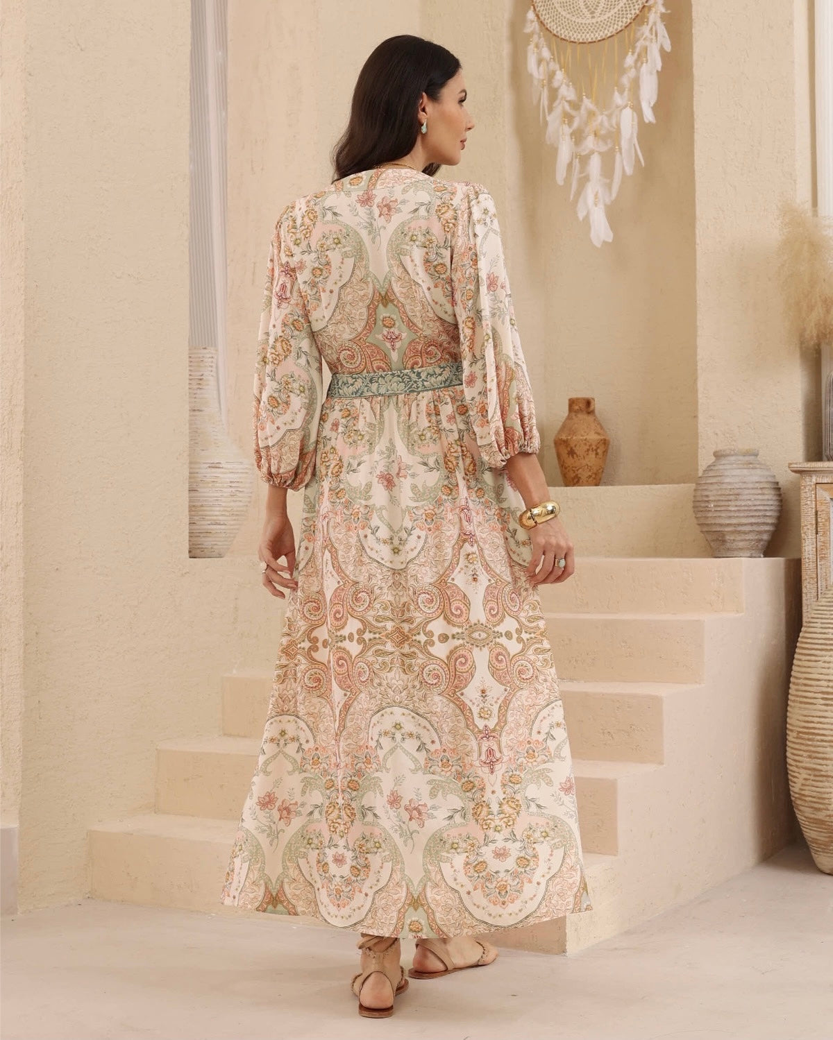 A woman viewed from behind wearing a long bohemian-style maxi dress with a beige, pink, and green paisley pattern, standing in front of beige stone steps.