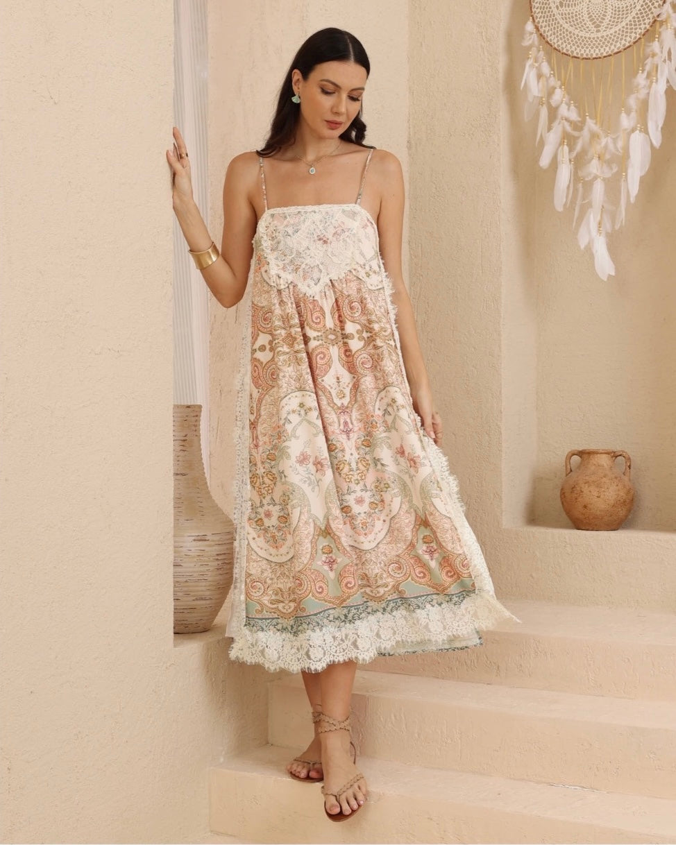 A woman models a bohemian-style midi dress with intricate paisley and floral embroidery in soft peach, beige, and sage green, featuring lace trim and spaghetti straps, set against a warm beige stucco wall and stone steps.
