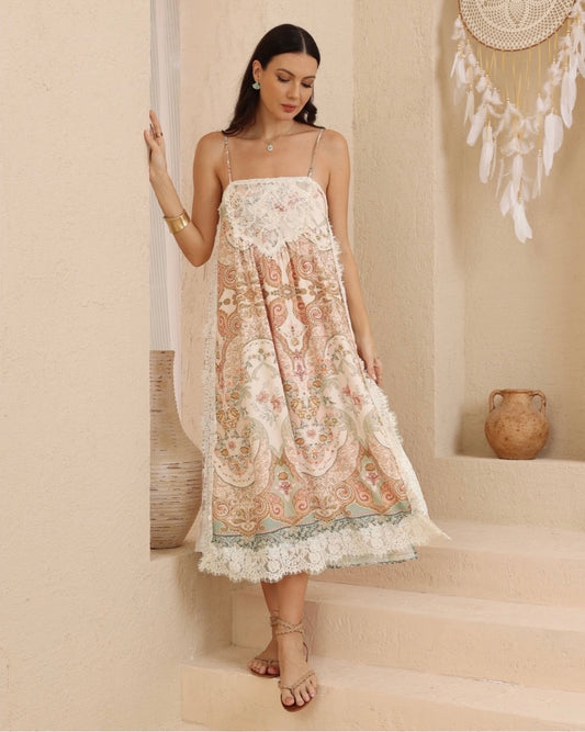 A woman models a bohemian-style midi dress with intricate paisley and floral embroidery in soft peach, beige, and sage green, featuring lace trim and spaghetti straps, set against a warm beige stucco wall and stone steps.