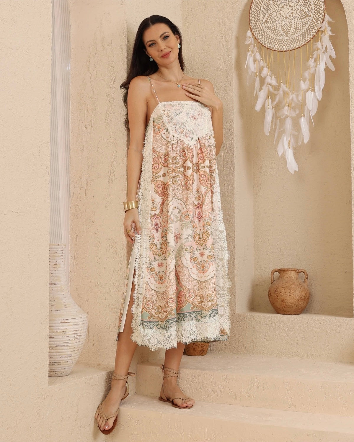 A woman models a bohemian-style midi dress with a cream base and intricate multicolor paisley and floral patterns. The dress features thin straps, and a lace trim hem, styled with neutral sandals in a warm, textured beige room decorated with a dreamcatcher and ceramic pots