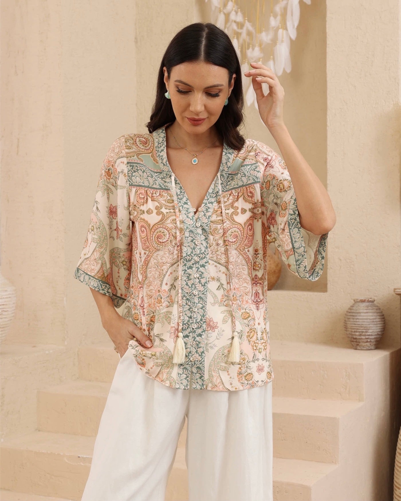 A woman models a short-sleeve V-neck blouse with a multicolor paisley and floral print in beige, green, and pink tones, paired with white wide-leg pants, set against a warm beige architectural background.