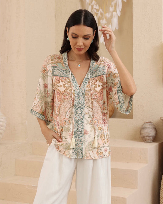 A woman models a short-sleeve V-neck blouse with a multicolor paisley and floral print in beige, green, and pink tones, paired with white wide-leg pants, set against a warm beige architectural background.