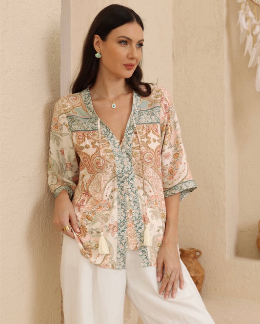 A woman models a bohemian-style printed blouse with three-quarter sleeves, a V-neckline, and tassel ties, paired with white pants in a warm, textured indoor setting.