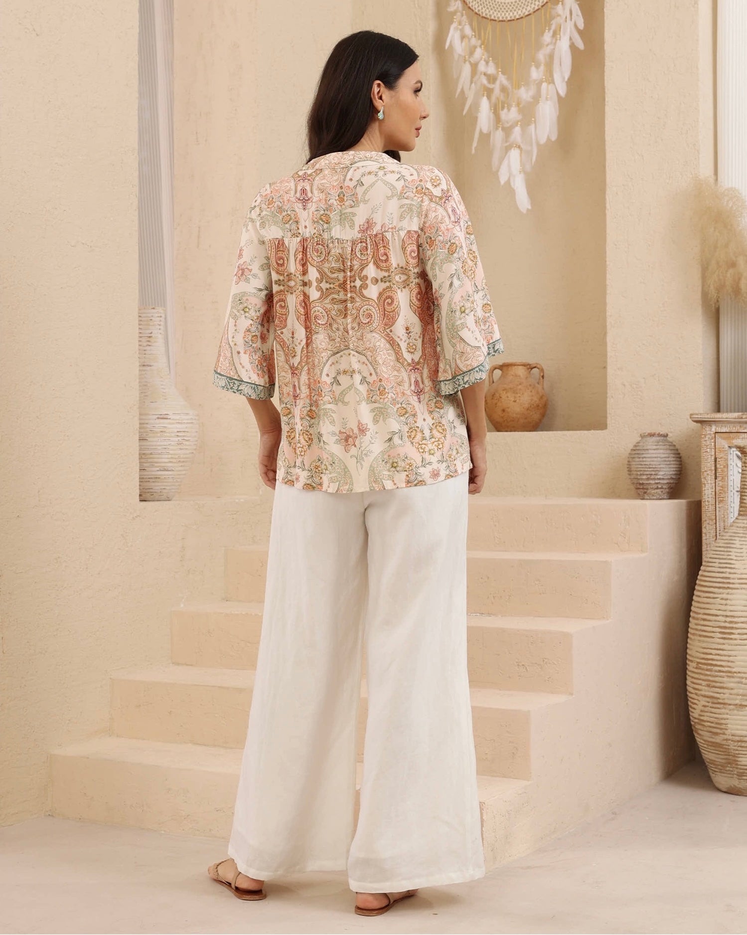 A woman stands with her back to the camera wearing a beige and pink paisley printed top with three-quarter sleeves and white wide-leg trousers, set against a warm beige architectural background with decorative vases and a dreamcatcher.