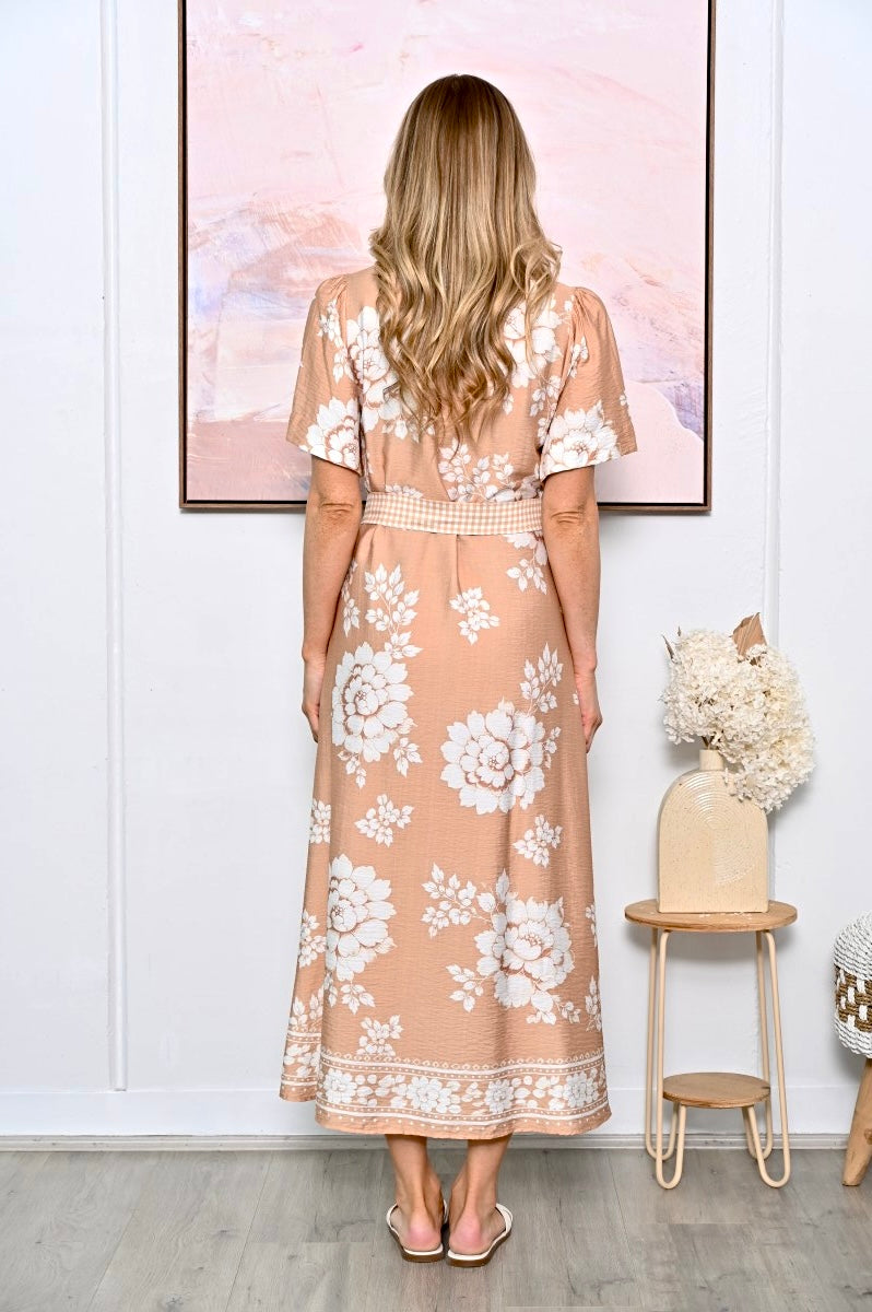 A woman stands with her back to the camera wearing a peach-colored maxi dress with a white floral pattern and a matching belt. She is in a room with a white paneled wall, a framed abstract print, and a small wooden side table with dried flowers.