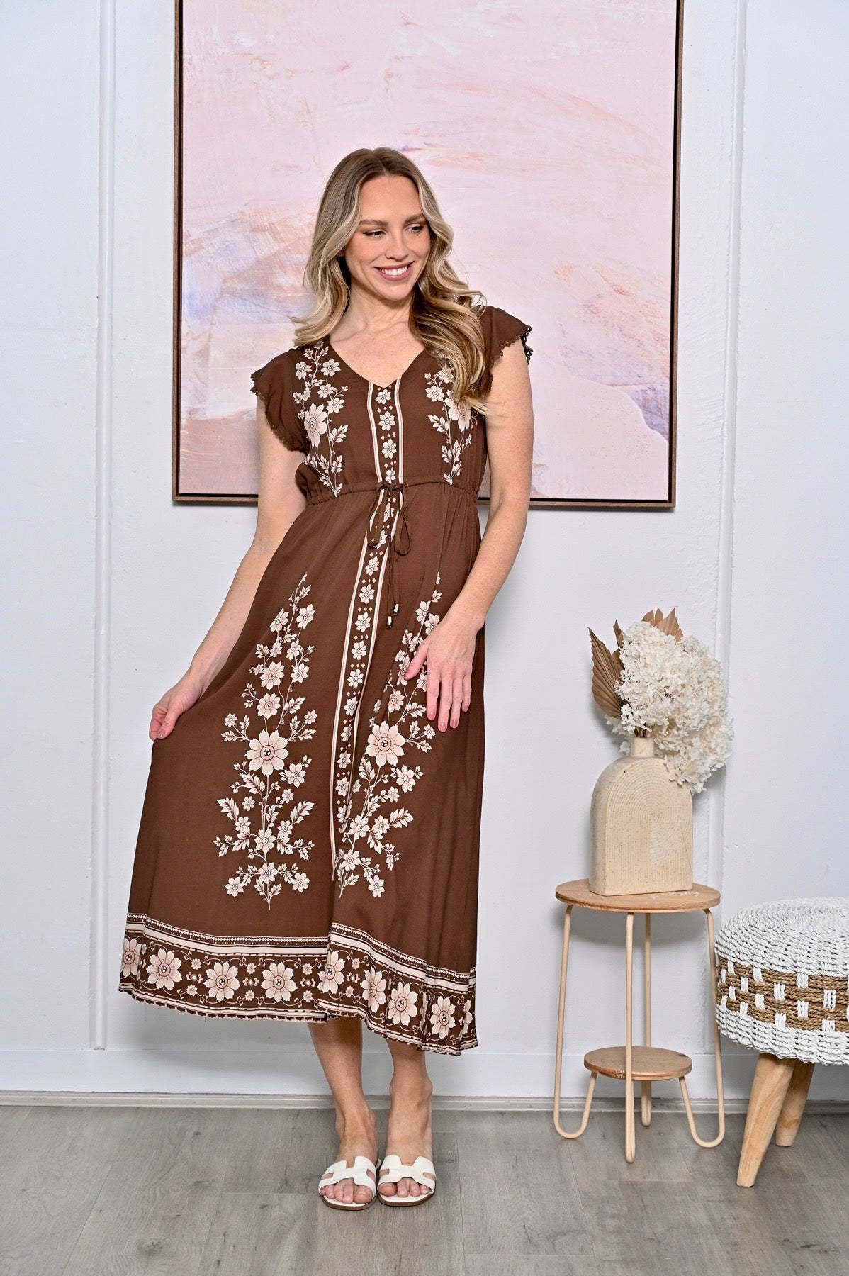 A woman modeling a brown midi dress with cream floral embroidery and a tie-front detail, standing in a styled room with a framed print, side table, and decorative vase.